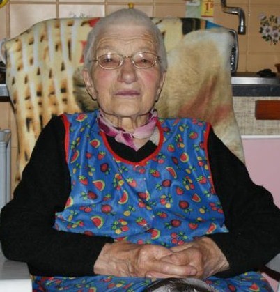 Starsza kobieta w okularach i fartuszku pozuje do zdjęcia.