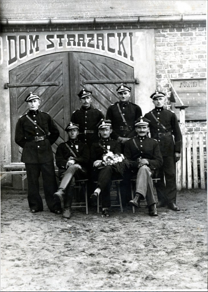 Siedmiu mężczyzn w mundurach strażackich pozuje do zdjęcia przed Domem Strażackim. A.A. Molski z bukietem kwiatów na kolanach.
