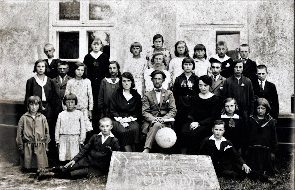 Na zdjęciu grupa dzieci w odświętnych strojach, pozująca do fotografii z trójką dorosłych, siedzących na krzesłach. Mężczyzna to A.A. Molski, jedną z kobiet - Maria Molska. 