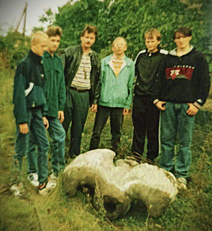 Grupa uczniów wraz z nauczycielami na tle zielonych krzewów i drzew. U ich stóp leżą fragmenty starego pomnika